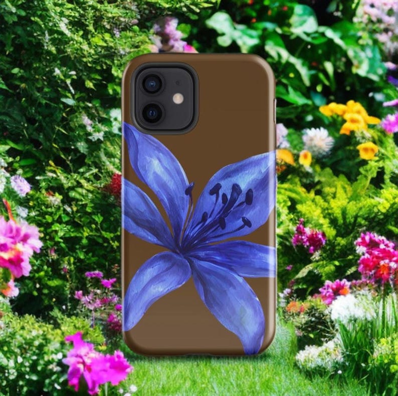 A brown phone case featuring a blue lily painting is positioned upright amidst a vibrant garden of blooming flowers and green foliage.