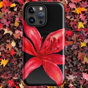 A black phone case with a red lily flower design rests on a bed of colorful autumn leaves.