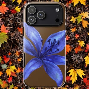 A brown phone case featuring a blue lily design is positioned vertically over a ground covered in fallen autumn leaves.