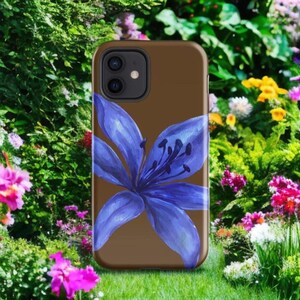A brown phone case with a large blue lily graphic is displayed vertically against a lush green garden filled with colorful flowers.