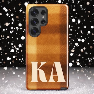 Puede incluir: Una funda de teléfono con un patrón de cuadros en tonos marrones y beige. La funda presenta las letras "KA" en crema en la parte inferior. La funda del teléfono tiene un módulo de cámara con múltiples lentes.