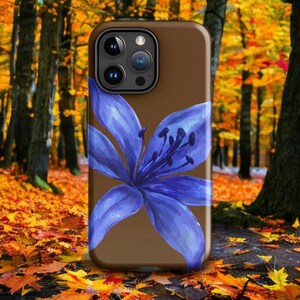 A brown phone case with a blue lily illustration is displayed upright in front of a forest with autumn foliage.