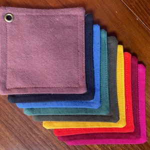 以下が含まれることがあります： 10色の布製のコースターのセット。それぞれ異なる色で、柔らかく、質感のある布地で作られています。上部の角に穴が開いており、吊り下げることができます。