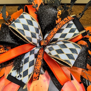 May include: A Halloween-themed decorative bow featuring a pumpkin shape. The bow has a gray and black diamond pattern with gold trim, accented by orange and black ribbons and glittery black elements. The pumpkin is orange with black cutouts.