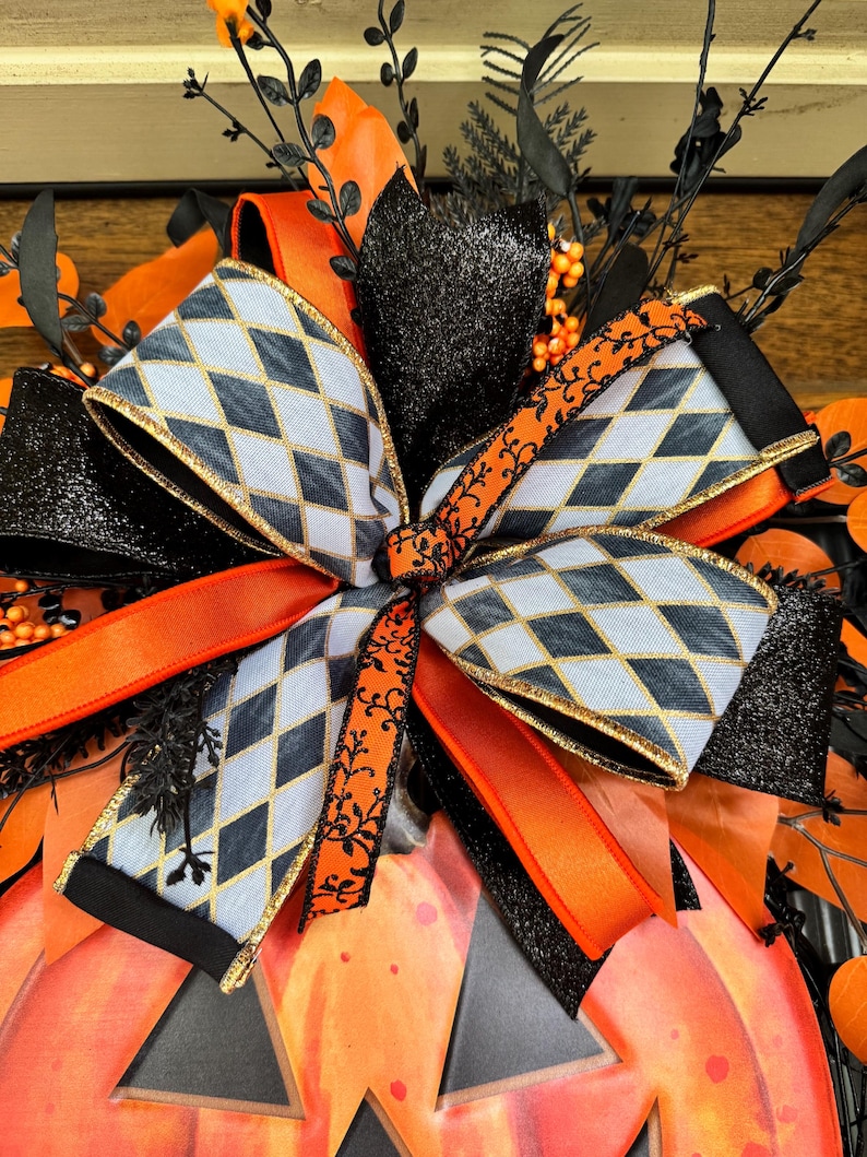 May include: A Halloween-themed decorative bow featuring a pumpkin shape. The bow has a gray and black diamond pattern with gold trim, accented by orange and black ribbons and glittery black elements. The pumpkin is orange with black cutouts.