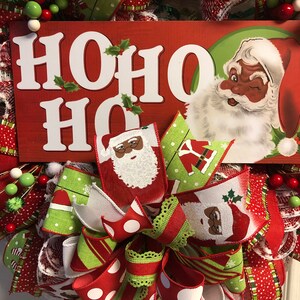 Puede incluir: Una corona de Navidad con un esquema de color rojo, blanco y verde. La corona presenta un letrero que dice "HO HO HO" y un Pap&aacute; Noel con la cara negra. La corona est&aacute; decorada con un lazo y otros adornos festivos.