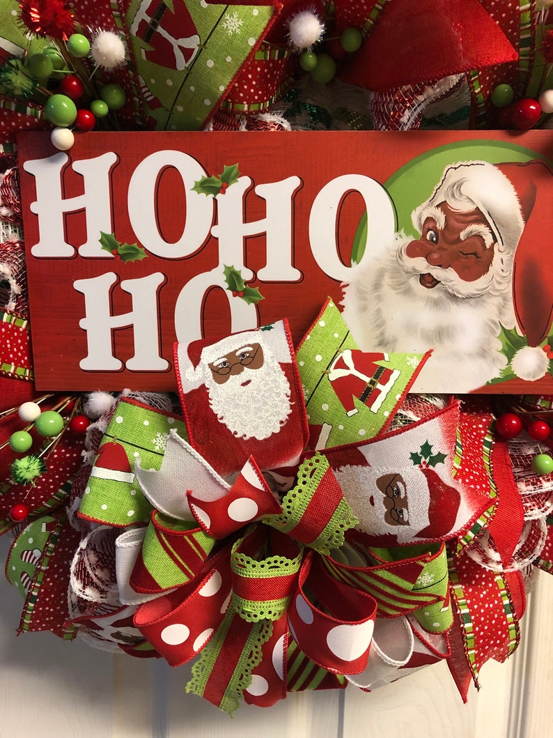 Puede incluir: Una corona de Navidad con un esquema de color rojo, blanco y verde. La corona presenta un letrero que dice "HO HO HO" y un Pap&aacute; Noel con la cara negra. La corona est&aacute; decorada con un lazo y otros adornos festivos.