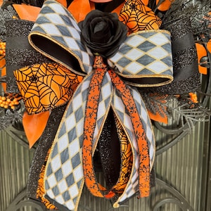 May include: A Halloween wreath with a large bow featuring a gray and white diamond pattern, orange spiderwebs, and a black rose. The wreath includes orange leaves and black accents, creating a festive and spooky decoration.