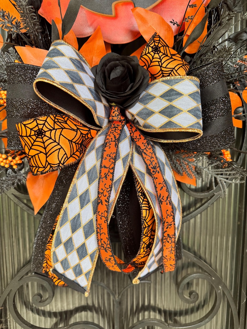 May include: A Halloween wreath with a large bow featuring a gray and white diamond pattern, orange spiderwebs, and a black rose. The wreath includes orange leaves and black accents, creating a festive and spooky decoration.
