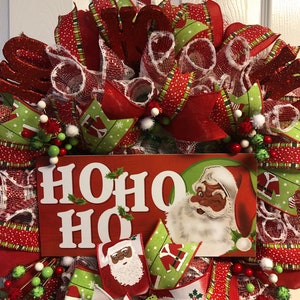 Puede incluir: Una corona de Navidad con cinta roja y verde, malla blanca y un letrero que dice "HOHO HO". El letrero presenta a un Pap&aacute; Noel negro con barba blanca y gorro rojo.