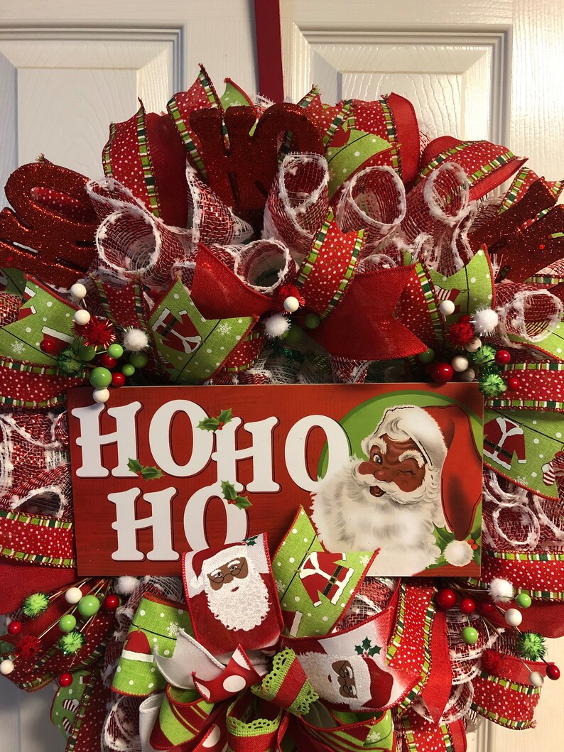Puede incluir: Una corona de Navidad con cinta roja y verde, malla blanca y un letrero que dice "HOHO HO". El letrero presenta a un Pap&aacute; Noel negro con barba blanca y gorro rojo.