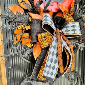 May include: A Halloween wreath featuring a large orange pumpkin with a carved face. The wreath is decorated with black and orange ribbons, black leaves, and a black rose. The ribbons have spiderweb and diamond patterns.