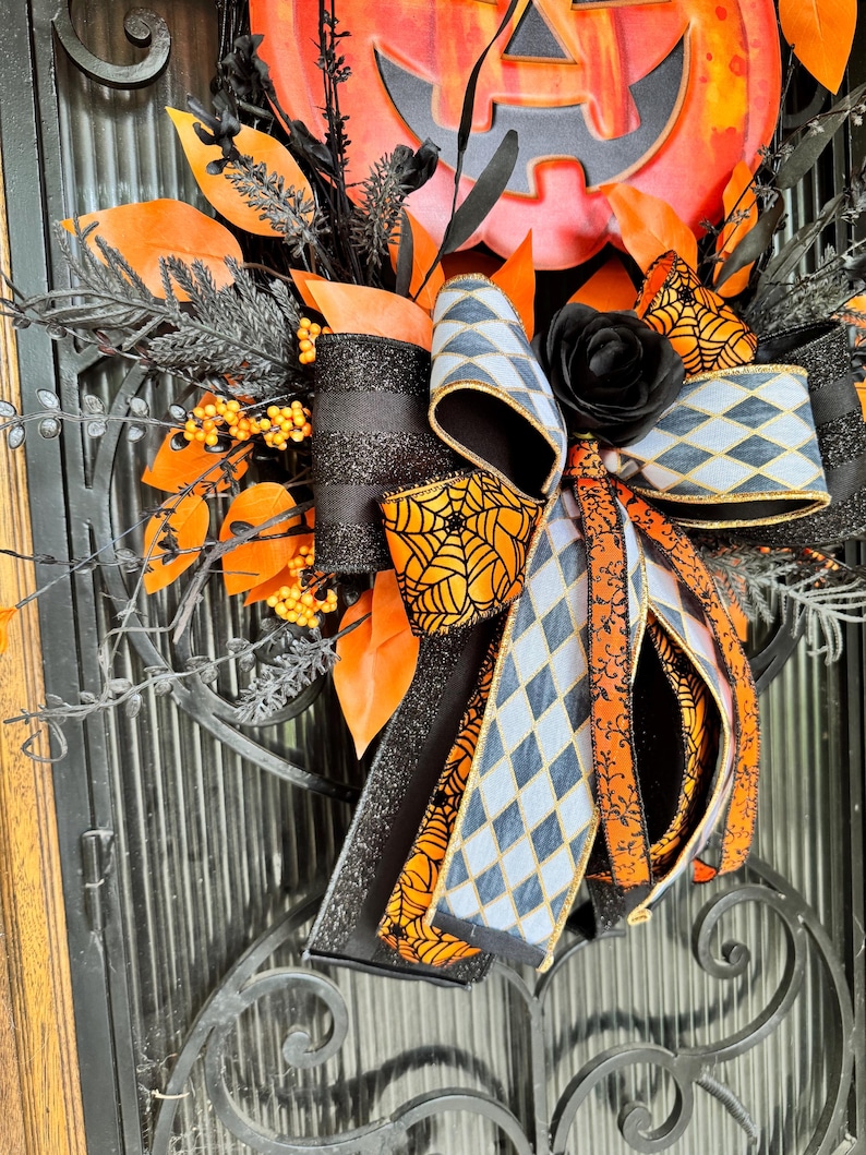 May include: A Halloween wreath featuring a large orange pumpkin with a carved face. The wreath is decorated with black and orange ribbons, black leaves, and a black rose. The ribbons have spiderweb and diamond patterns.