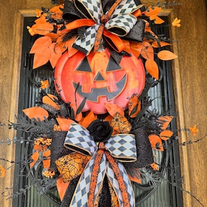 May include: A Halloween-themed wreath featuring a large orange pumpkin with a carved face. The wreath is adorned with black and orange ribbons, leaves, and decorative elements. The design incorporates a mix of textures and patterns, creating a festive and eye-catching display.