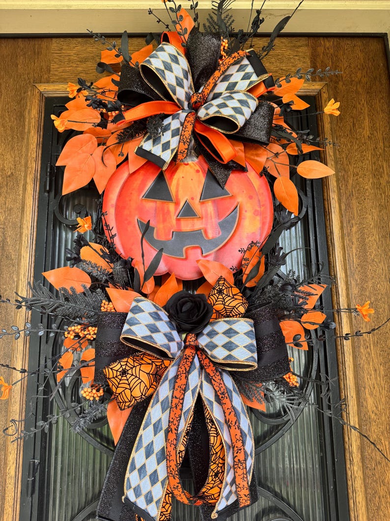 May include: A Halloween-themed wreath featuring a large orange pumpkin with a carved face. The wreath is adorned with black and orange ribbons, leaves, and decorative elements. The design incorporates a mix of textures and patterns, creating a festive and eye-catching display.