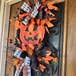 May include: A Halloween-themed door wreath featuring a large orange pumpkin with a carved face. The wreath is adorned with black and orange ribbons, black foliage, and orange leaf accents. The design includes a mix of textures and patterns for a festive look.