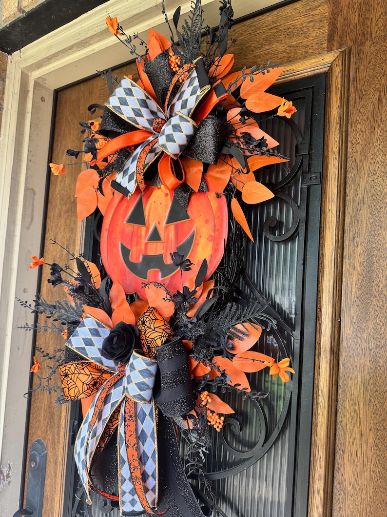 May include: A Halloween-themed door wreath featuring a large orange pumpkin with a carved face. The wreath is adorned with black and orange ribbons, black foliage, and orange leaf accents. The design includes a mix of textures and patterns for a festive look.