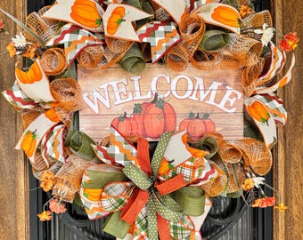 May include: A fall-themed wreath with orange, green, and cream ribbons, featuring pumpkin designs and the word "WELCOME" on a wooden sign. The wreath is adorned with artificial flowers and is displayed on a dark door.