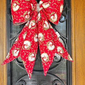 May include: A large fabric bow in a vibrant red color featuring a repeating pattern of Santa Claus faces surrounded by green holly wreaths. The bow has two wide top loops and two long, pointed tails hanging down, with a small plaid center knot.