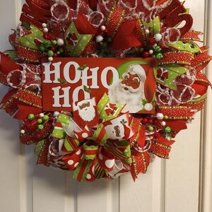 Puede incluir: Una corona de Navidad roja, verde y blanca con un letrero de "HO HO HO" que presenta a un Pap&aacute; Noel de dibujos animados. La corona est&aacute; decorada con cinta de lunares rojos y blancos, cinta de rayas verdes y blancas y malla blanca.
