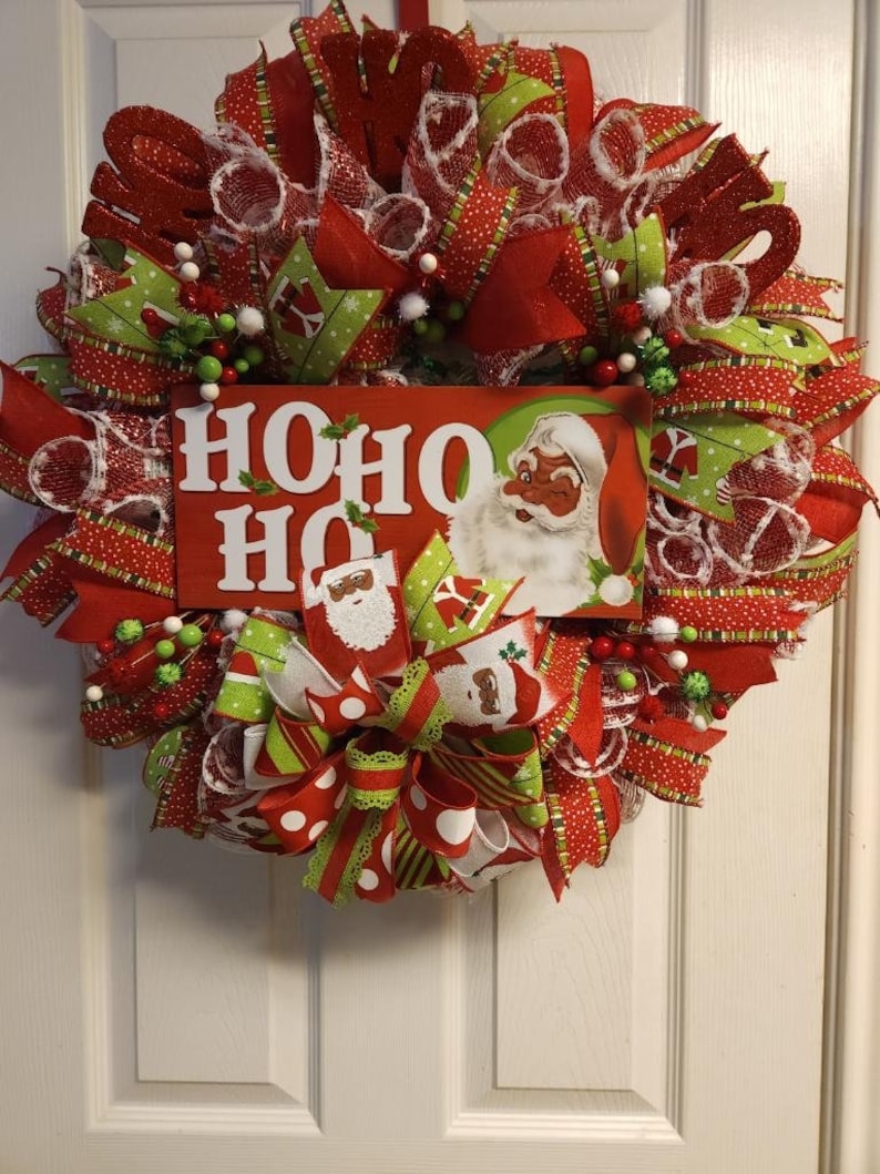 Puede incluir: Una corona de Navidad roja, verde y blanca con un letrero de "HO HO HO" que presenta a un Pap&aacute; Noel de dibujos animados. La corona est&aacute; decorada con cinta de lunares rojos y blancos, cinta de rayas verdes y blancas y malla blanca.