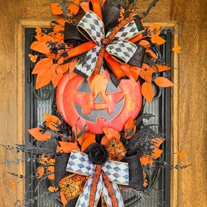 May include: A Halloween-themed wreath featuring a large orange jack-o'-lantern with a smiling face. The wreath is adorned with orange and black ribbons, leaves, and decorative elements. The design includes a black rose and a bow with a diamond pattern.