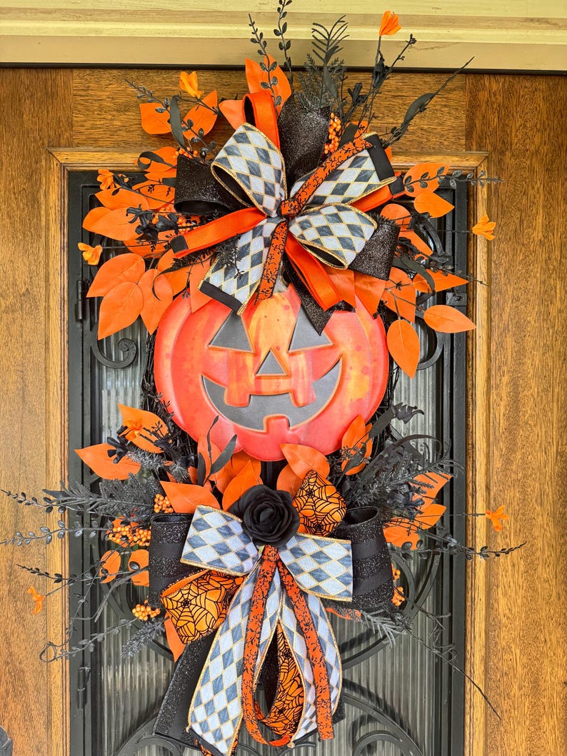 May include: A Halloween-themed wreath featuring a large orange jack-o'-lantern with a smiling face. The wreath is adorned with orange and black ribbons, leaves, and decorative elements. The design includes a black rose and a bow with a diamond pattern.
