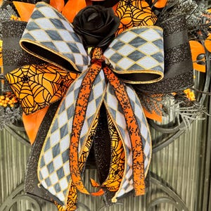 May include: A Halloween-themed wreath with a black rose, orange and black ribbons, and decorative elements. The bow features a gray and white diamond pattern with gold trim, and orange spiderweb accents. The wreath is set against a dark background.