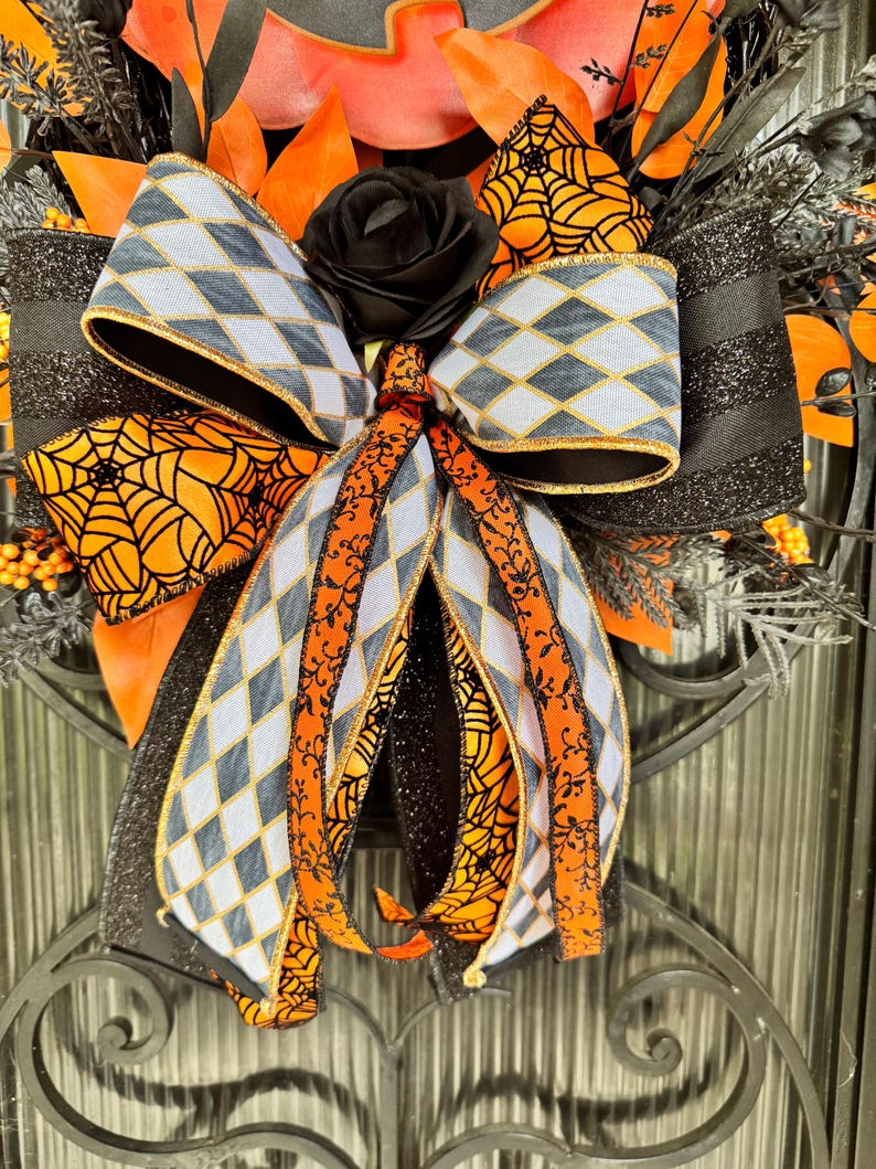 May include: A Halloween-themed wreath with a black rose, orange and black ribbons, and decorative elements. The bow features a gray and white diamond pattern with gold trim, and orange spiderweb accents. The wreath is set against a dark background.