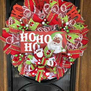 Puede incluir: Una corona de Navidad con cinta roja y verde, malla blanca y un letrero que dice "HOHO HO". El letrero presenta a un Pap&aacute; Noel de dibujos animados con la cara negra.