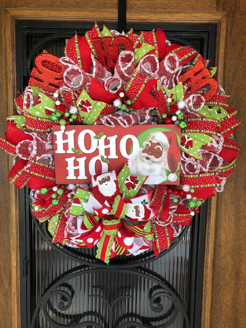 Puede incluir: Una corona de Navidad con cinta roja y verde, malla blanca y un letrero que dice "HOHO HO". El letrero presenta a un Pap&aacute; Noel de dibujos animados con la cara negra.
