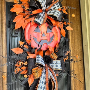 May include: A Halloween-themed wreath featuring a large orange pumpkin with a carved face. The wreath is adorned with black and orange ribbons, leaves, and decorative elements. The design includes a black rose and a bow with a spiderweb pattern.