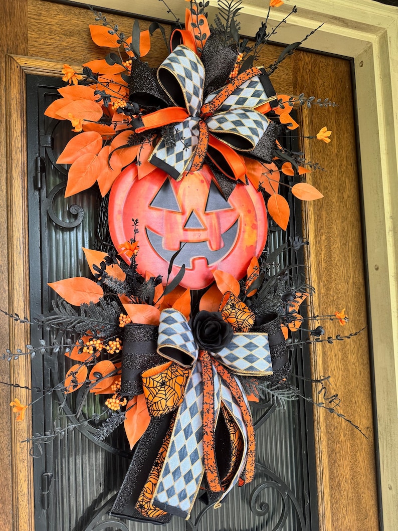 May include: A Halloween-themed wreath featuring a large orange pumpkin with a carved face. The wreath is adorned with black and orange ribbons, leaves, and decorative elements. The design includes a black rose and a bow with a spiderweb pattern.