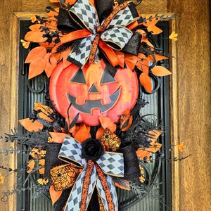 May include: A Halloween-themed wreath featuring a smiling jack-o'-lantern centerpiece. The wreath is adorned with orange and black ribbons, leaves, and decorative elements. The design includes a black rose and diamond-patterned bows.