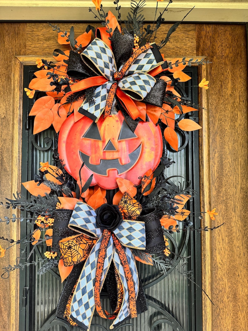 May include: A Halloween-themed wreath featuring a smiling jack-o'-lantern centerpiece. The wreath is adorned with orange and black ribbons, leaves, and decorative elements. The design includes a black rose and diamond-patterned bows.