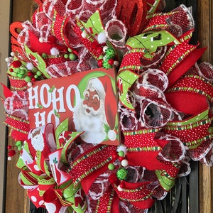 Puede incluir: Una corona de Navidad roja, verde y blanca con un letrero que dice "HO HO HO" y una imagen de Santa Claus. La corona est&aacute; decorada con cinta de lunares rojos y blancos, cinta de rayas verdes y blancas y malla blanca.
