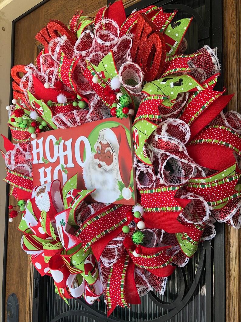 Puede incluir: Una corona de Navidad roja, verde y blanca con un letrero que dice "HO HO HO" y una imagen de Santa Claus. La corona est&aacute; decorada con cinta de lunares rojos y blancos, cinta de rayas verdes y blancas y malla blanca.
