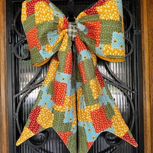 May include: A large decorative bow made of quilted fabric featuring a patchwork pattern of red, yellow, green, and light blue squares with floral and polka dot prints. The center of the bow is cinched with a dark green and white plaid fabric.