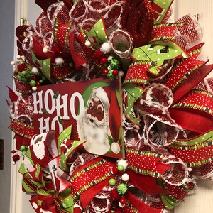 Puede incluir: Una corona de Navidad roja, verde y blanca con un letrero que dice "HO HO HO" y una imagen de Santa Claus. La corona est&aacute; decorada con cinta de lunares rojos y blancos, cinta de rayas verdes y blancas y malla blanca.