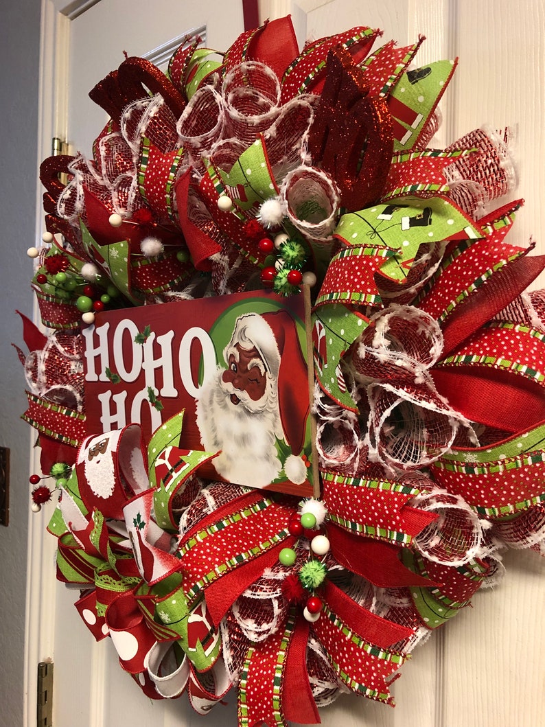 Puede incluir: Una corona de Navidad roja, verde y blanca con un letrero que dice "HO HO HO" y una imagen de Santa Claus. La corona est&aacute; decorada con cinta de lunares rojos y blancos, cinta de rayas verdes y blancas y malla blanca.