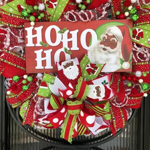 Puede incluir: Una corona de Navidad con cinta roja, verde y blanca, un letrero que dice "HO HO HO" y un Pap&aacute; Noel con la cara negra.