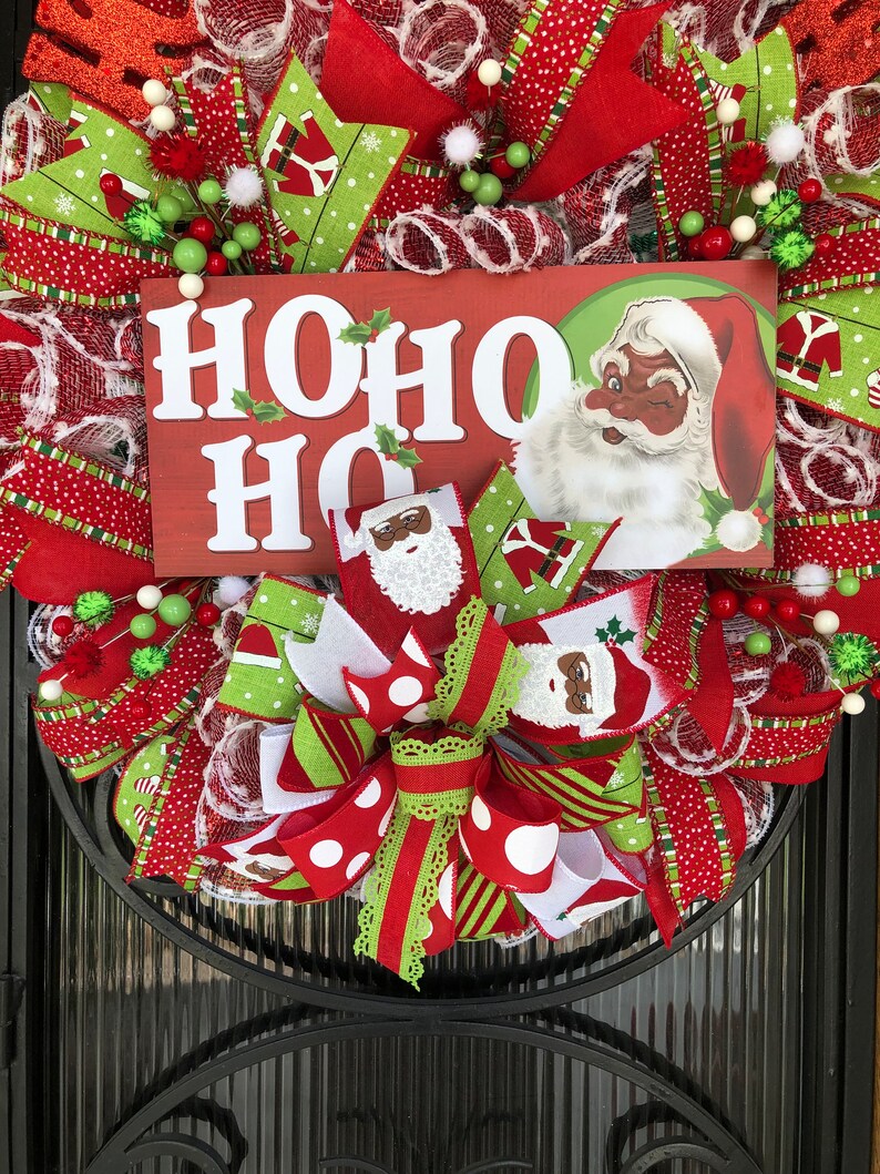 Puede incluir: Una corona de Navidad con cinta roja, verde y blanca, un letrero que dice "HO HO HO" y un Pap&aacute; Noel con la cara negra.