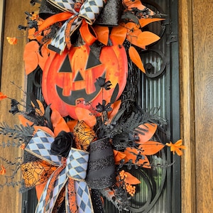 May include: A Halloween-themed wreath featuring a large orange pumpkin with a carved face. The wreath is adorned with black and orange ribbons, leaves, and decorative elements. The design incorporates a mix of textures and patterns, creating a festive and eye-catching display.