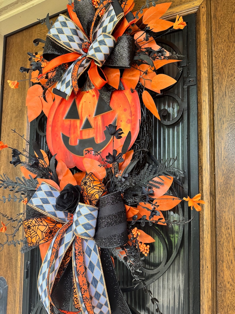 May include: A Halloween-themed wreath featuring a large orange pumpkin with a carved face. The wreath is adorned with black and orange ribbons, leaves, and decorative elements. The design incorporates a mix of textures and patterns, creating a festive and eye-catching display.