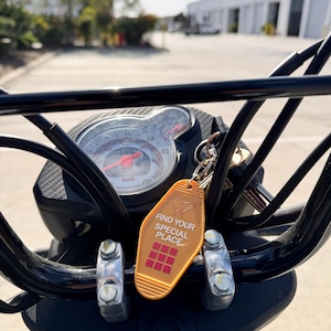 May include: A gold motel key chain with the text "Find Your Special Place" and a red and white grid pattern. The key chain is attached to a black motorcycle handlebar.