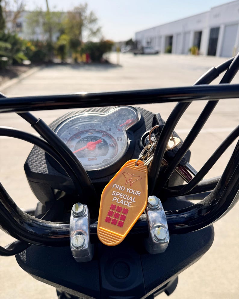May include: A gold motel key chain with the text "Find Your Special Place" and a red and white grid pattern. The key chain is attached to a black motorcycle handlebar.