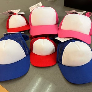 Puede incluir: Seis gorras de camionero en blanco en varios colores, incluyendo azul, rojo y rosa. Las gorras tienen una parte trasera de malla blanca y un panel frontal blanco.