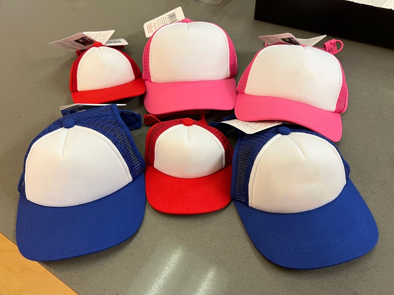 Puede incluir: Seis gorras de camionero en blanco en varios colores, incluyendo azul, rojo y rosa. Las gorras tienen una parte trasera de malla blanca y un panel frontal blanco.