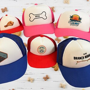 Puede incluir: Cinco gorras de camionero diferentes con dise&ntilde;os variados. Las gorras son blancas con una parte trasera de malla y tienen viseras de diferentes colores. Los dise&ntilde;os incluyen una cordillera monta&ntilde;osa, un hueso con la palabra "Mochi" en &eacute;l, una palmera con las palabras "Summer Vibes", un barco con las palabras "Wild Adventures" y una rama con las palabras "Soy el gerente de la sucursal".
