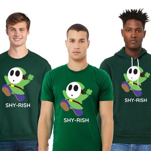 May include: Three people wearing green shirts with a cartoon character wearing a mask and holding a shamrock. The shirts say "Shy-Rish".